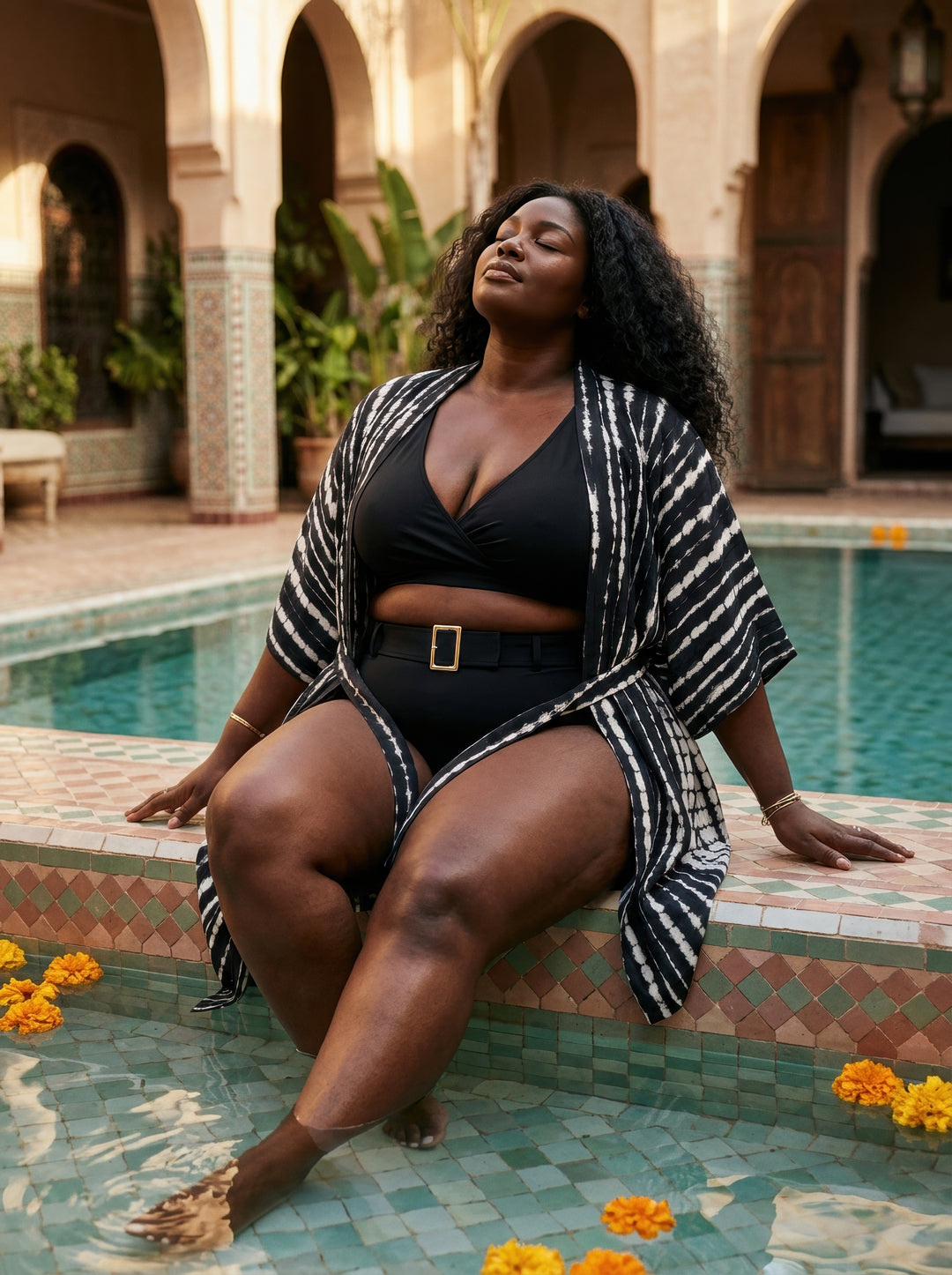 Woman in a black swimsuit and striped cover-up sitting by a pool with decorative tiles and flowers.