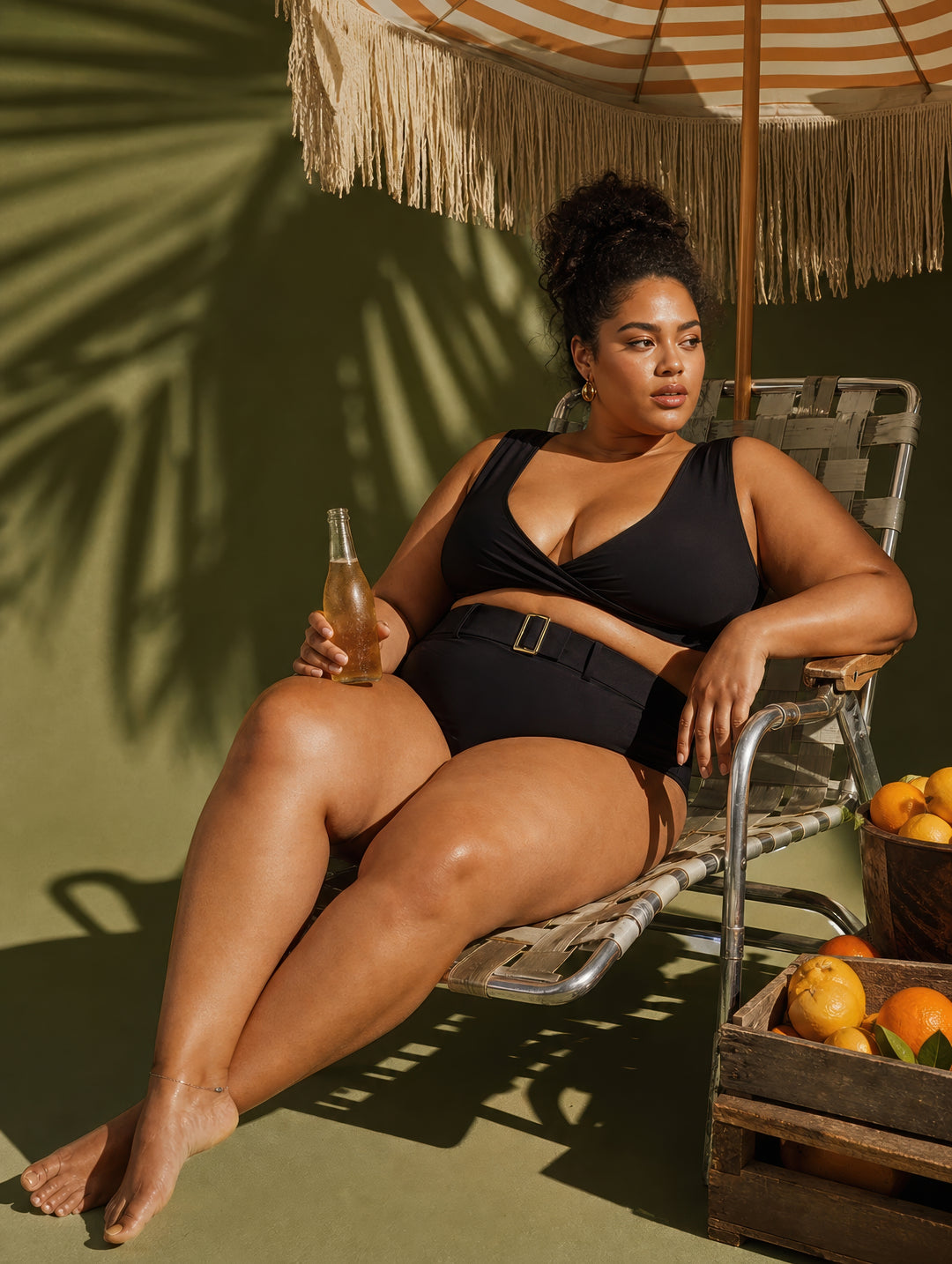 Woman in black swimsuit sitting under a striped umbrella with a bottle in a tropical setting