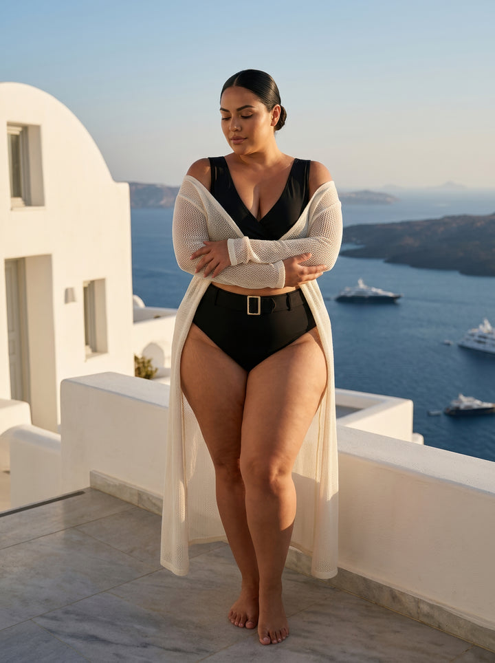 Woman in a black swimsuit with a white cover-up standing on a balcony with a scenic ocean view.